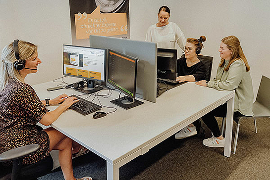 Vier Frauen mit Headsets sitzen sich am Schreibtisch gegenüber