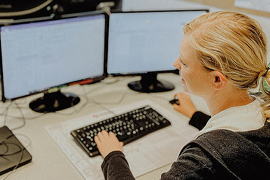 Blick auf zwei Bildschirme, von hinten ist eine Frau erkennbar, die am PC arbeitet